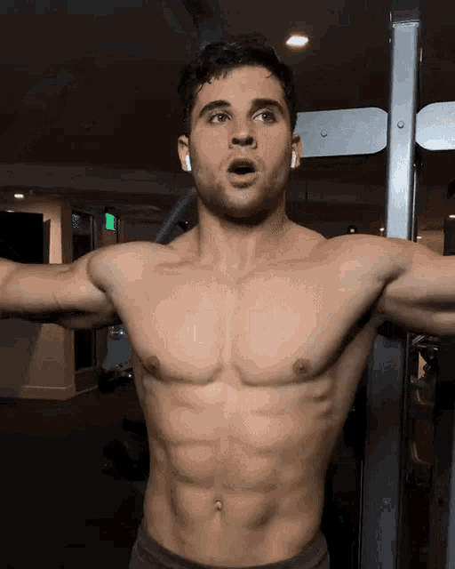 a shirtless man wearing ear buds stands in a gym with his mouth open