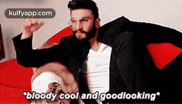 a man with a beard is sitting on a red couch with a dog and says `` bloody cool and goodlooking '' .