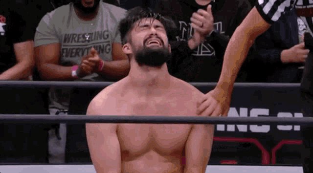 a man with a beard is kneeling down in a wrestling ring while a referee looks on .