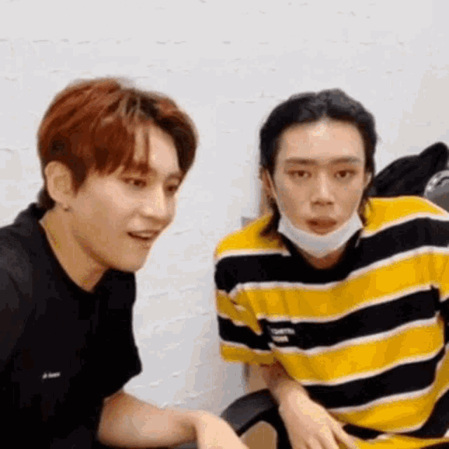 two young men wearing masks are sitting next to each other . one of the men is wearing a yellow and black striped shirt .
