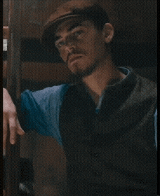a man wearing a hat and vest is leaning against a wooden wall