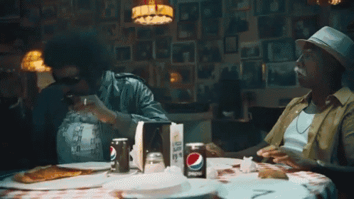 a can of pepsi sits on a table with two men eating pizza
