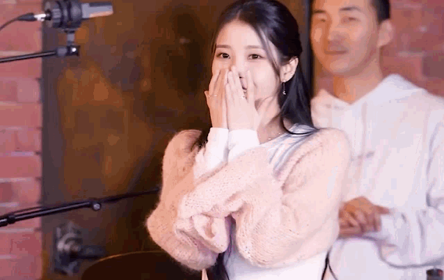 a woman in a pink sweater is covering her mouth with her hands while a man stands behind her .