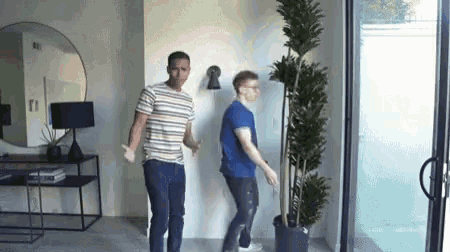 two men are standing next to a potted plant in a room