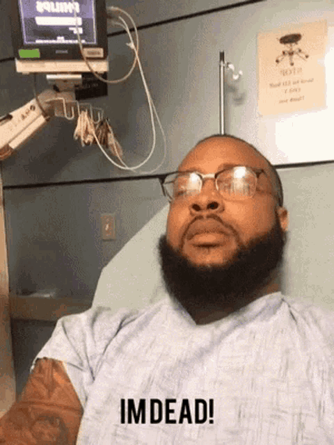 a man with glasses and a beard is laying in a hospital bed with the words im dead above him