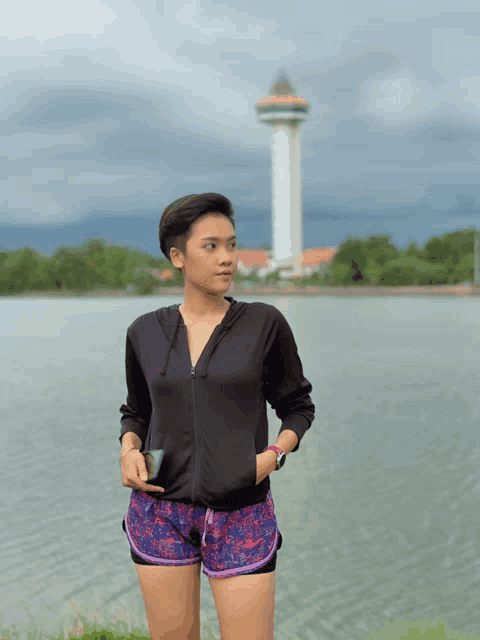 a woman in shorts and a black jacket stands in front of a lake