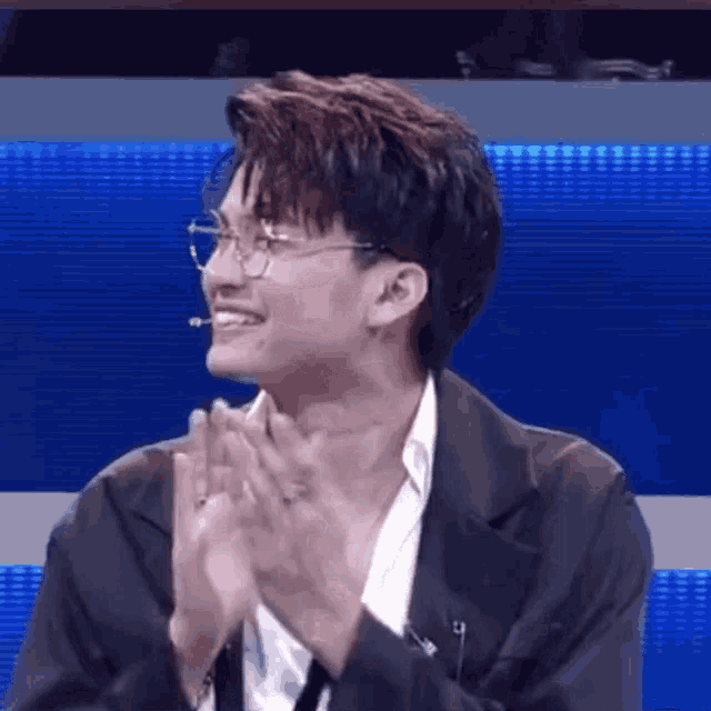 a man wearing glasses is clapping his hands while sitting in front of a blue screen .
