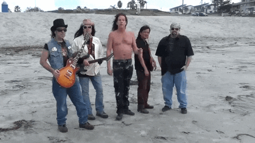 a group of men standing on a beach with one man holding an orange guitar