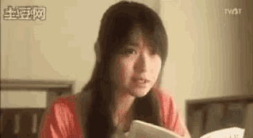 a woman is sitting at a table reading a book and making a funny face .