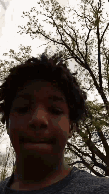 a young man with dreadlocks is standing in front of a tree and looking at the camera .