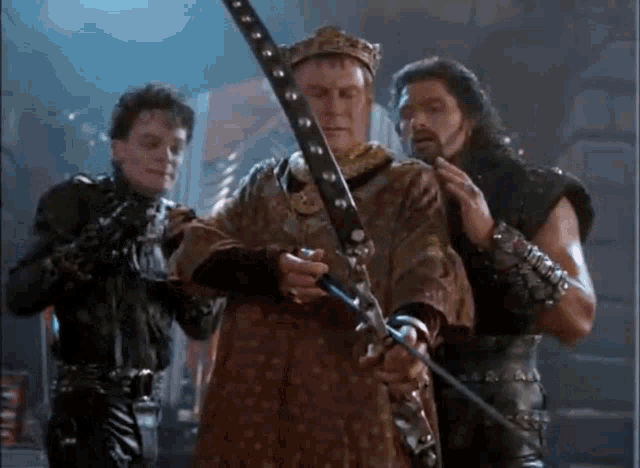 a man in a crown is holding a sword in front of two other men in leather armor .
