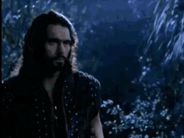 a man with long hair and a beard is standing in the woods holding a sword and a glowing object .