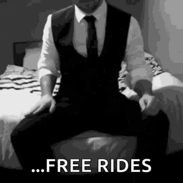 a man in a suit and tie is sitting on a bed with his legs crossed and says `` free rides '' .