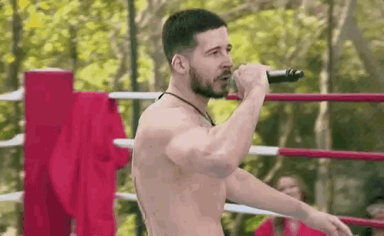 a shirtless man is standing in a boxing ring singing into a microphone .