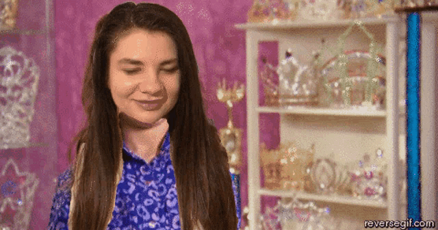 a woman in a blue shirt is standing in front of a shelf full of tiaras .