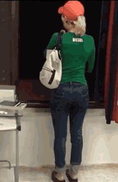 a woman in a green shirt and blue jeans is standing in front of a window holding a white bag .