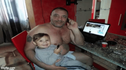 a shirtless man is holding a small child and talking on a cell phone