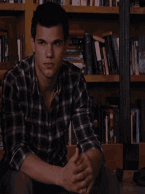 a man wearing a plaid shirt sits in front of a bookshelf