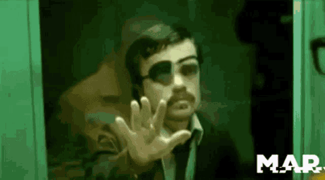 a man with a mustache and sunglasses is standing in front of a green door with his hand out .