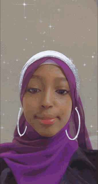 a woman wearing a purple hijab and hoop earrings is wearing a red tag that says right now