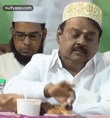 a man wearing a white hat and glasses is sitting at a table with another man .