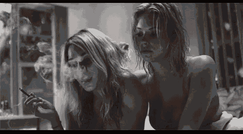 two naked women are smoking cigarettes together in a black and white photo .