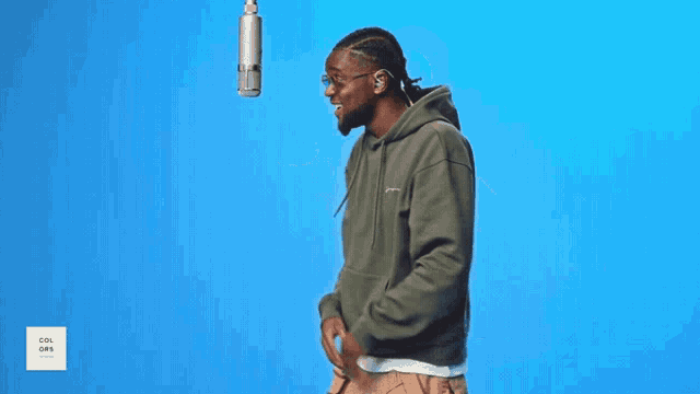 a man wearing glasses and a green hoodie stands in front of a blue background and a microphone