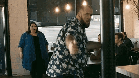 a man in a floral shirt is dancing in a restaurant while a woman stands behind him .
