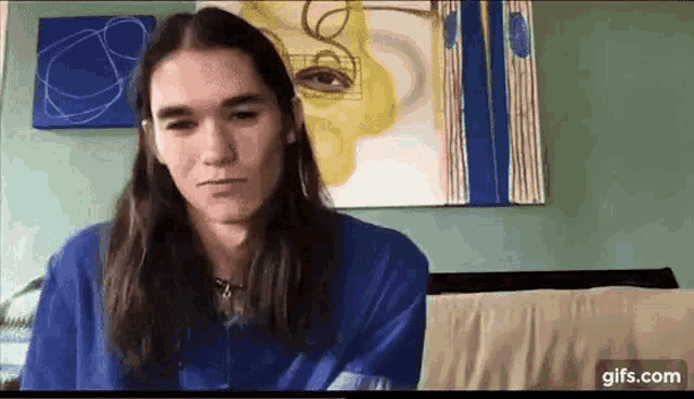 a young man with long hair is sitting on a couch in front of a painting of a face .
