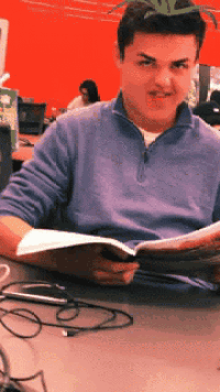 a man in a blue sweater is sitting at a desk reading a book
