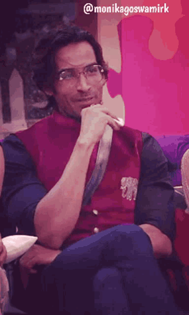 a man wearing glasses and a purple vest is sitting on a couch with his hands folded