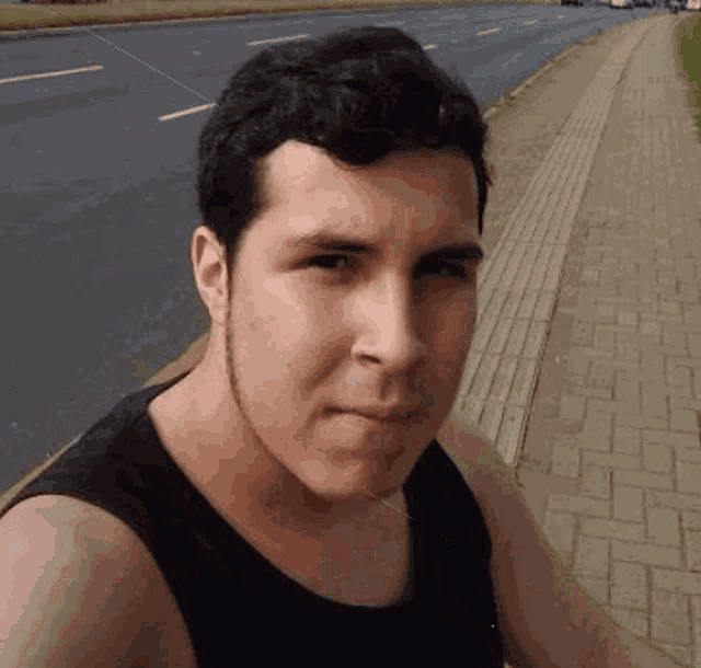 a man in a black tank top is standing on a sidewalk next to a street .
