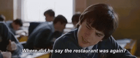 a boy is sitting at a desk in a classroom talking to another boy .