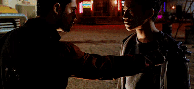 two men are standing next to each other in front of a building with neon lights .