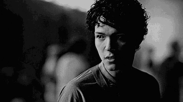 it is a black and white photo of a young man with curly hair looking at the camera .