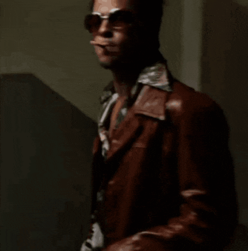 a man wearing sunglasses and a leather jacket is smoking a cigarette in a dark room .