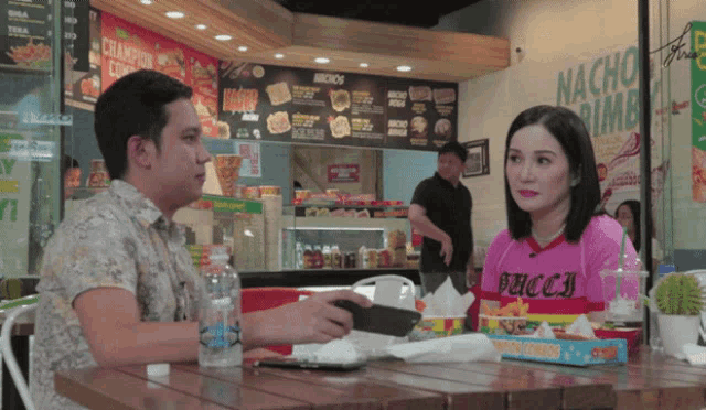 a man and a woman sit at a table in front of a sign that says nacho 's