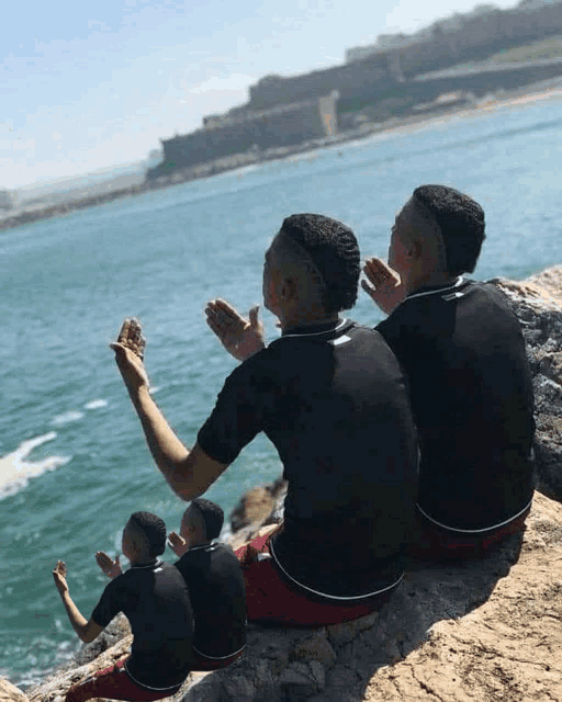 three men sit on a rock near the ocean with their hands in the air