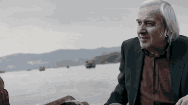 a man in a suit and red shirt is sitting by the water looking at the water .