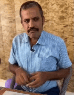 a man with a mustache is sitting at a table wearing a blue plaid shirt .
