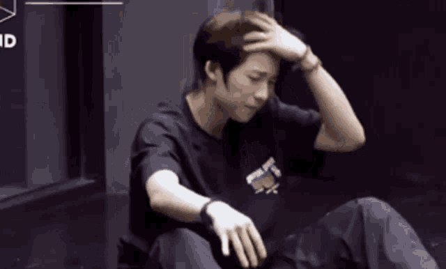 a man in a black shirt is sitting on the floor with his hand on his forehead .