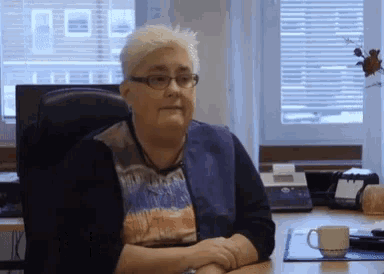 an older woman wearing glasses sits at a desk with her hands folded