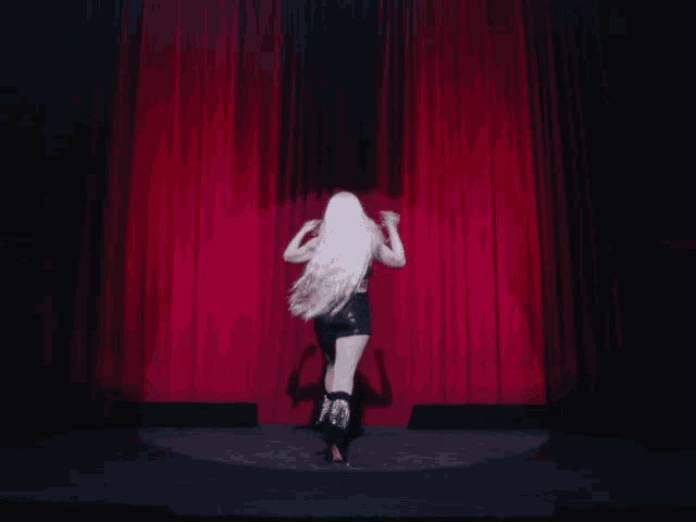 a woman with long white hair stands on a stage in front of a red curtain