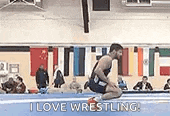 a man is kneeling on a wrestling mat in a gym while a crowd watches .