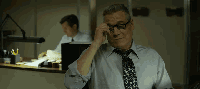 a man in a tie adjusts his glasses while another man looks on