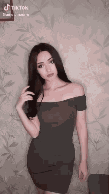 a woman in a grey off the shoulder dress stands in front of a wall with a tiktok watermark