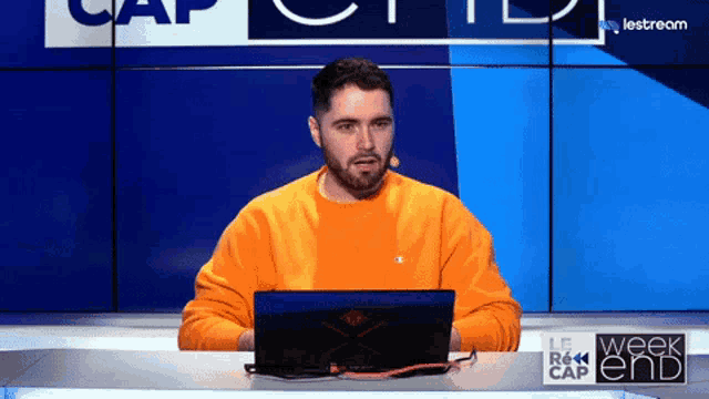 a man in an orange shirt is sitting at a desk with a laptop on it
