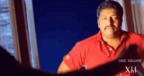 a man in a red shirt with a mustache is looking out a window .