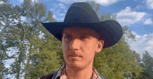 a man wearing a cowboy hat looks at the camera with trees in the background