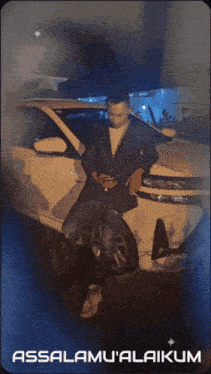 a man in a suit sits next to a white car with the words assalamu'alaikum on the bottom right